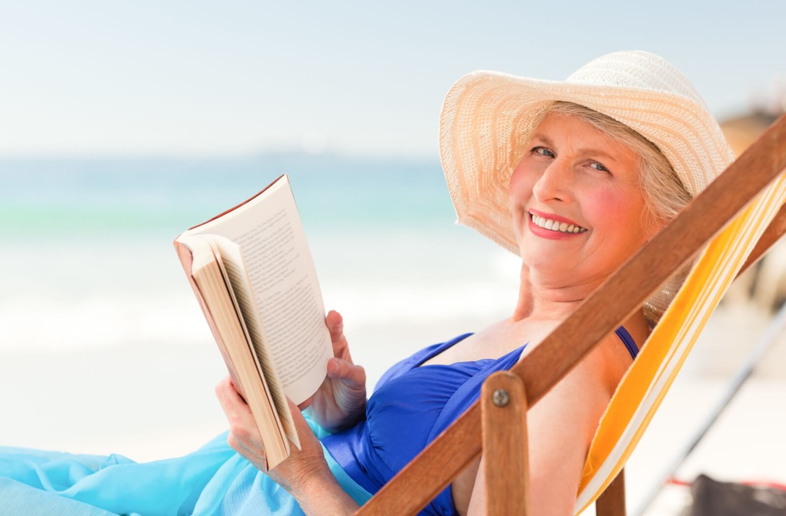 An older adult in a sun hat holds a book and smiles on the beach.