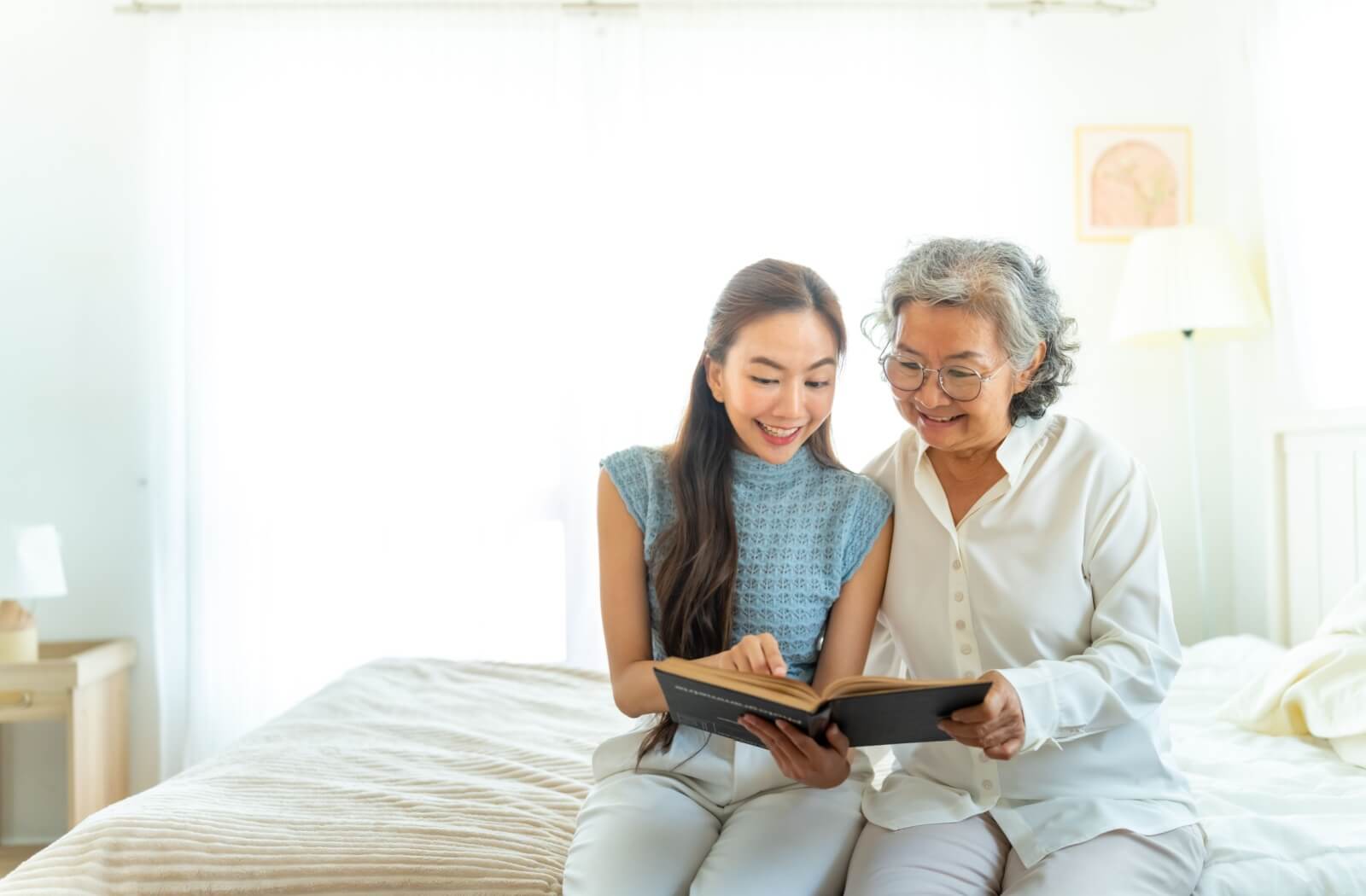 A young adult sits with their grandparent on the edge of a bed, reading a book out loud together