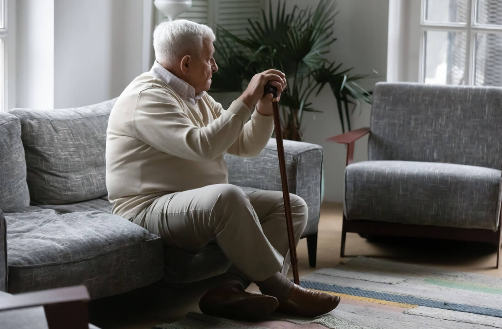 A senior with dementia holds onto their walking cane while staring sadly at an empty chair in a quiet, gray apartment