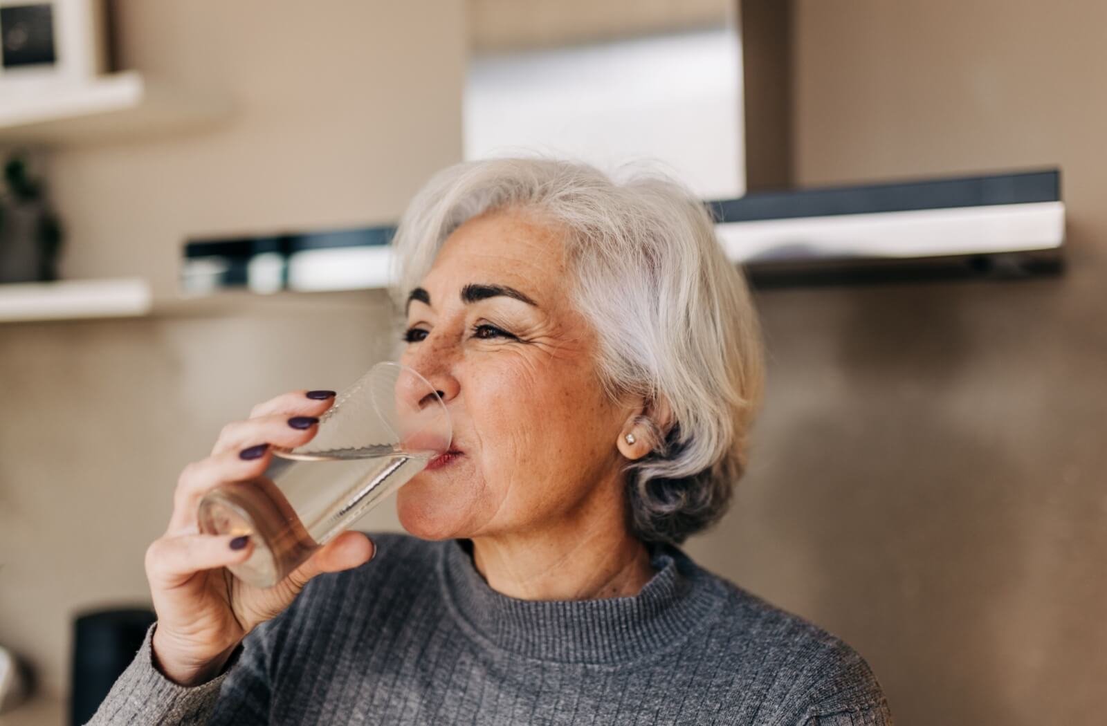 A senior with black fingernails happily drinks a glass of water first thing in the morning to promote all-day hydration