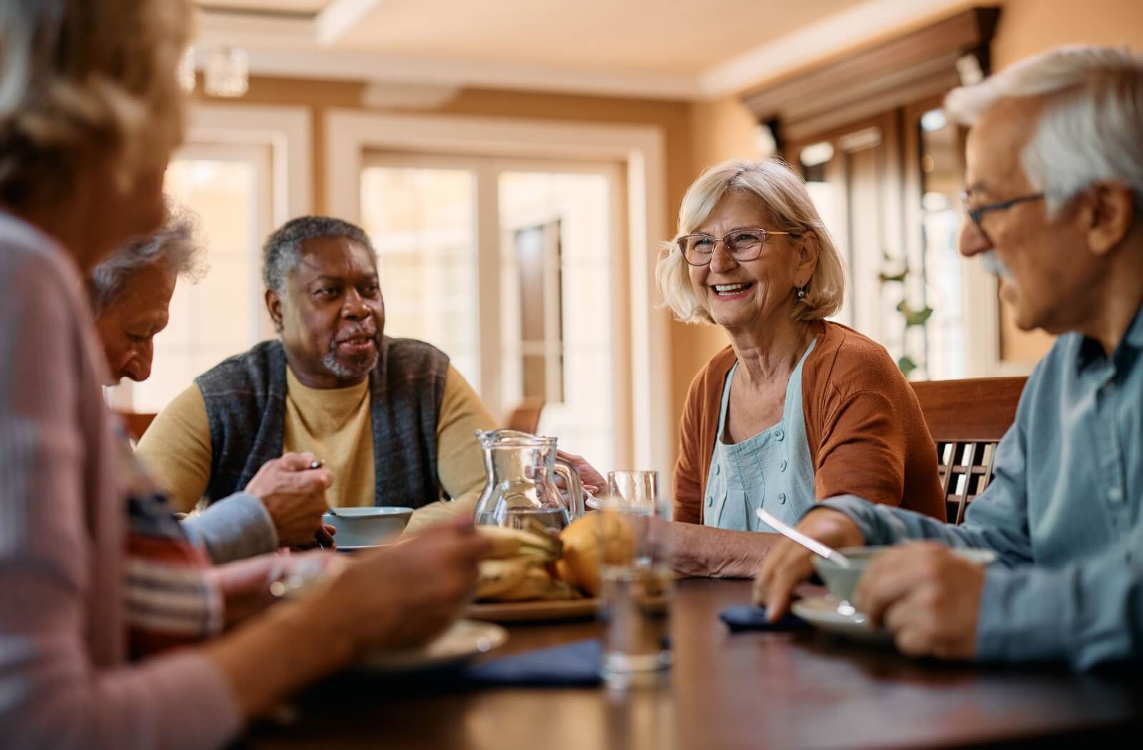 A group of senior friends laugh and talk while eating lunch in an assisted living community.