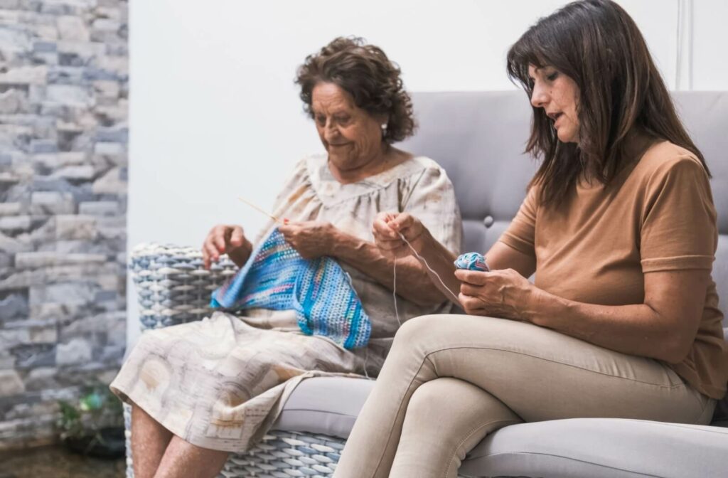 An adult opens a wool ball as they talk to their senior parent, who is knitting while sitting on a sofa in an assisted living community.