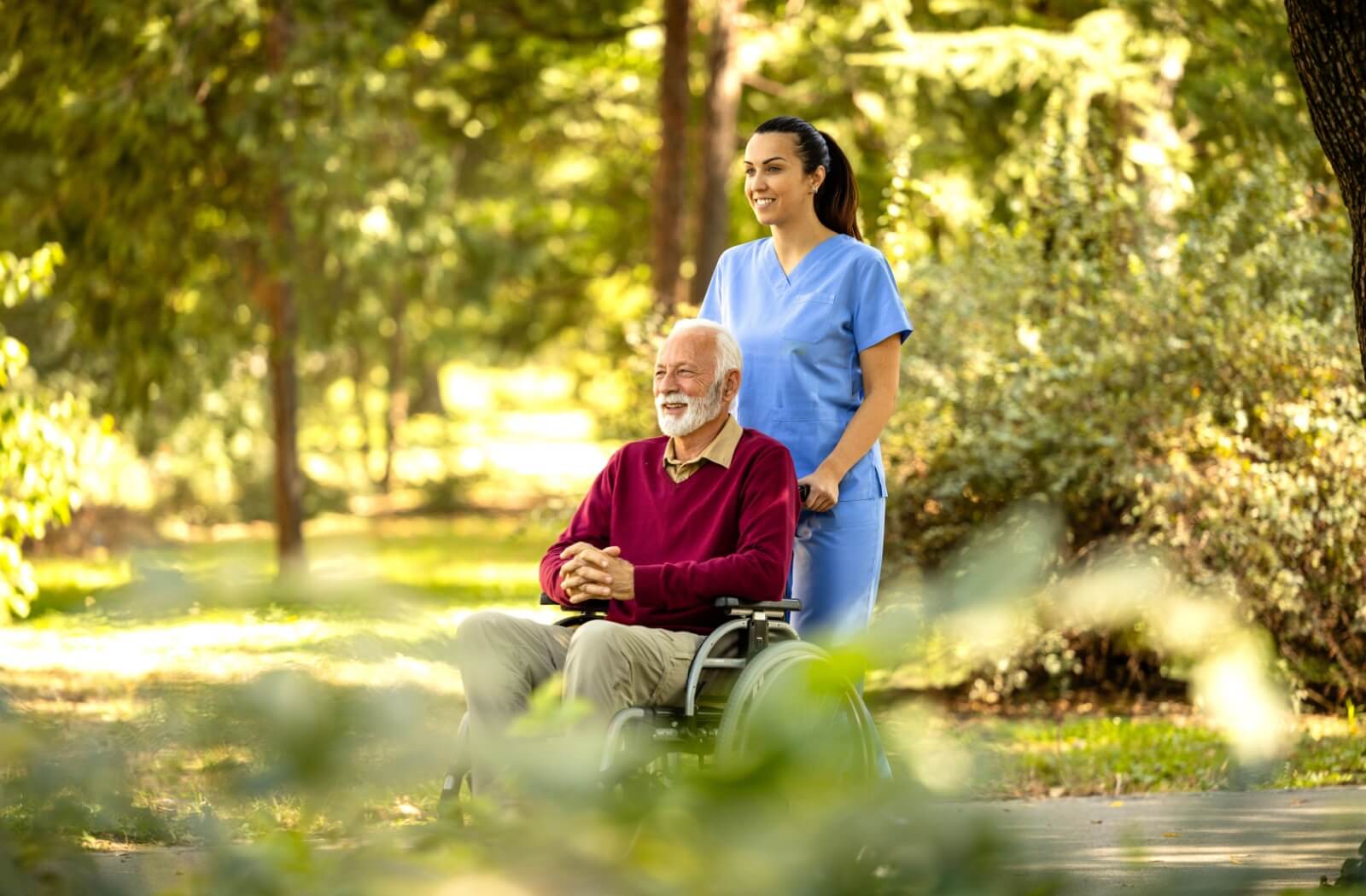 A senior being pushed through a park in their wheelchair by a nurse and smiling as they enjoy the time together.