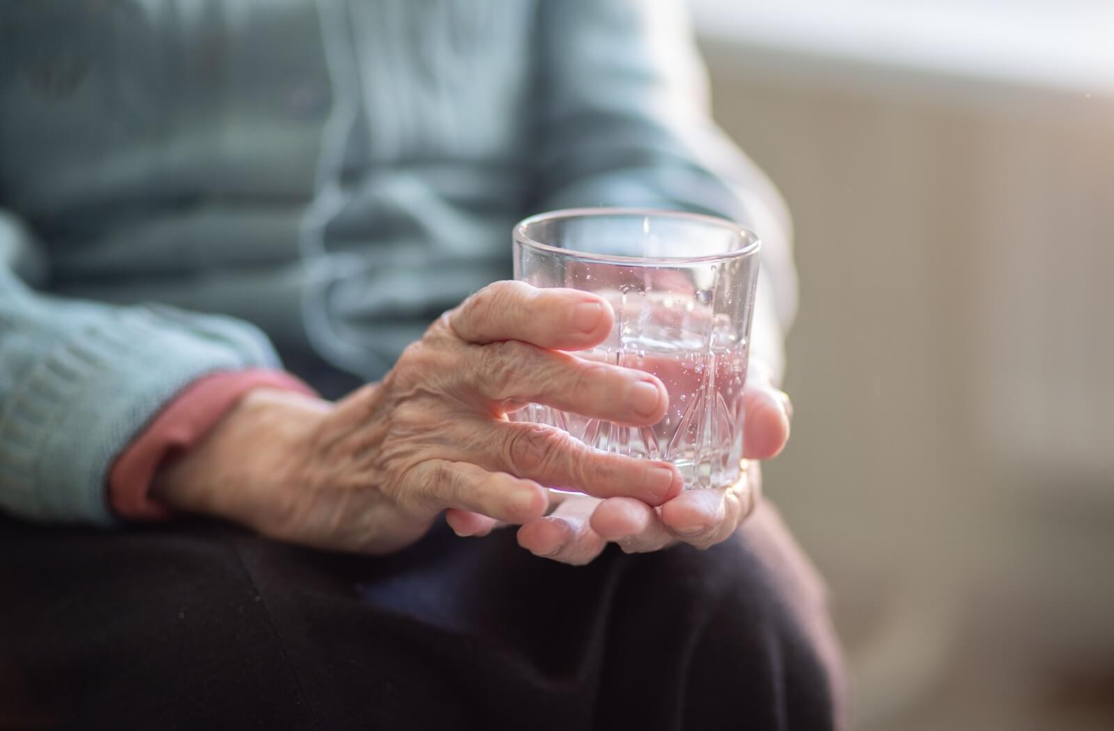 A close-up image an older adult holding a glass of water while they sit on their couch.