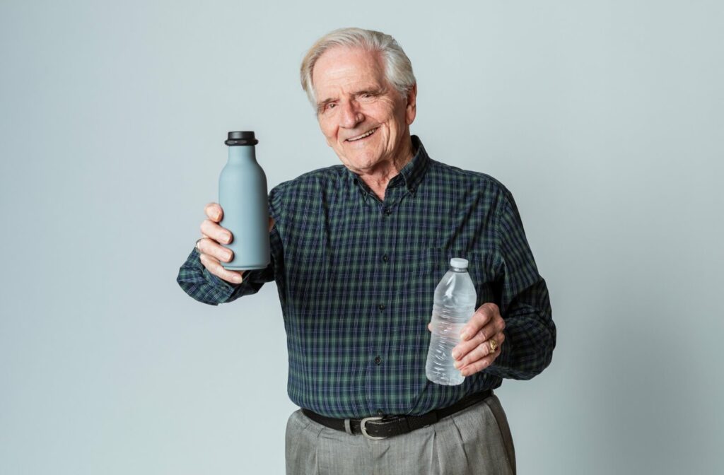 An older adult holding two bottles of water in front of a gray background, offering one to the camera.