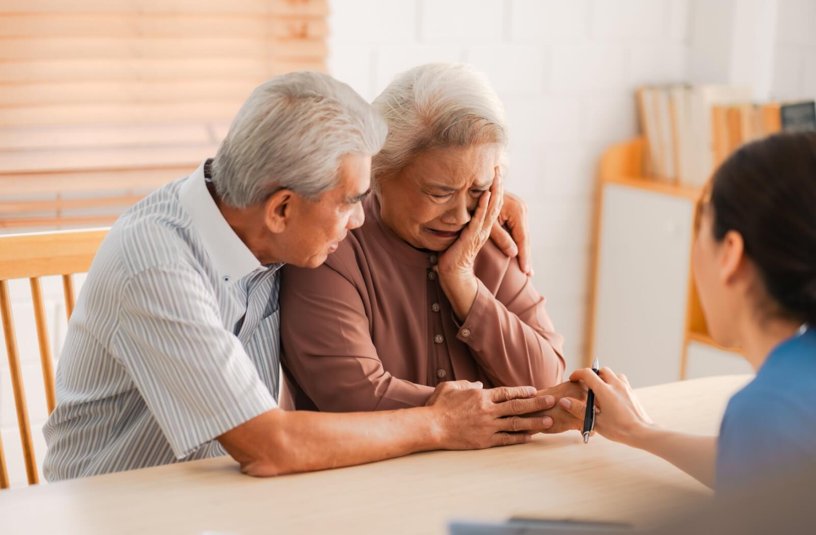 A senior comforts another emotional senior while a caregiver provides assistance at a table in a nursing home setting
