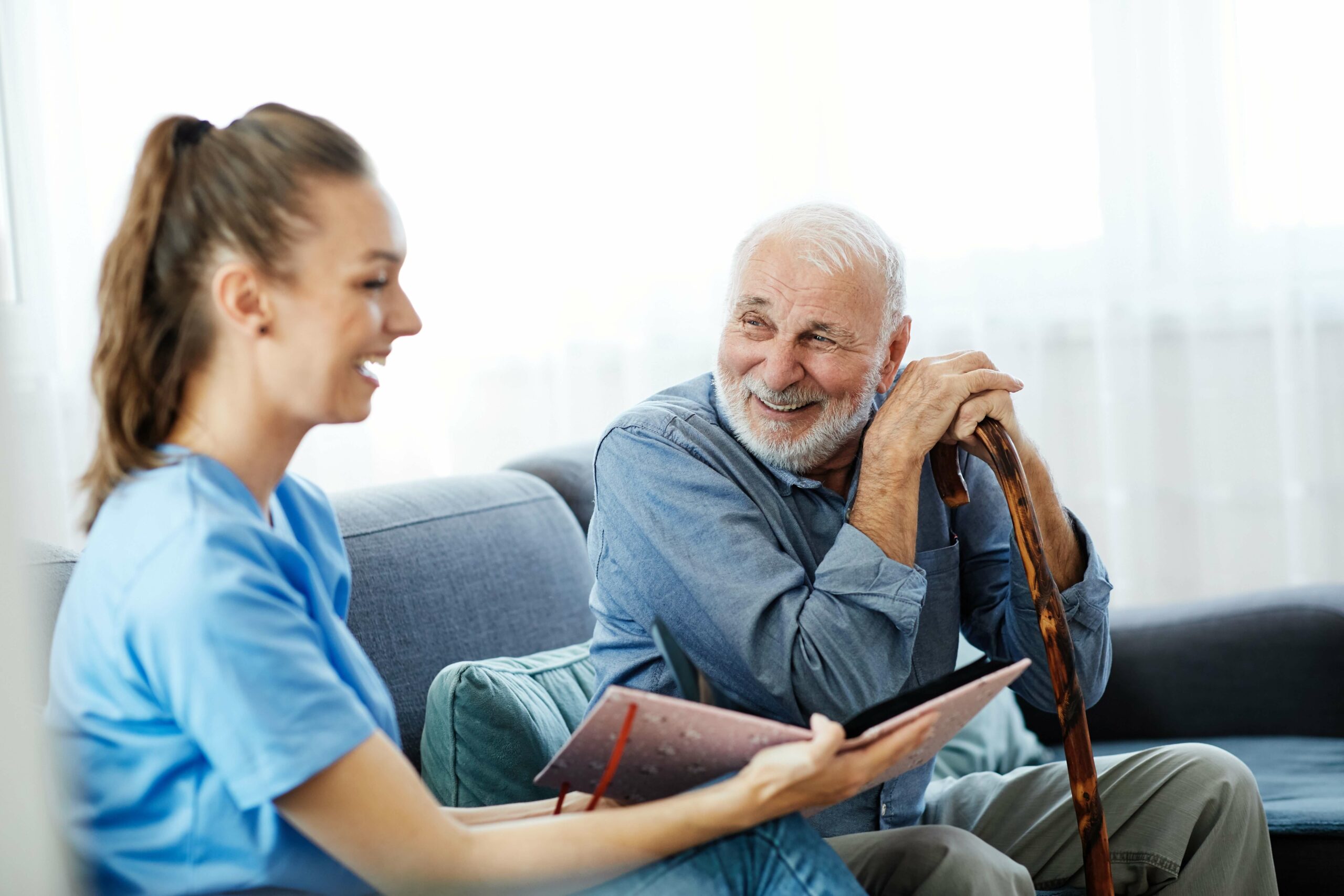 Nurse in blue scrubs and older man with cane smiling and laughing while looking through a photo album on a couch.