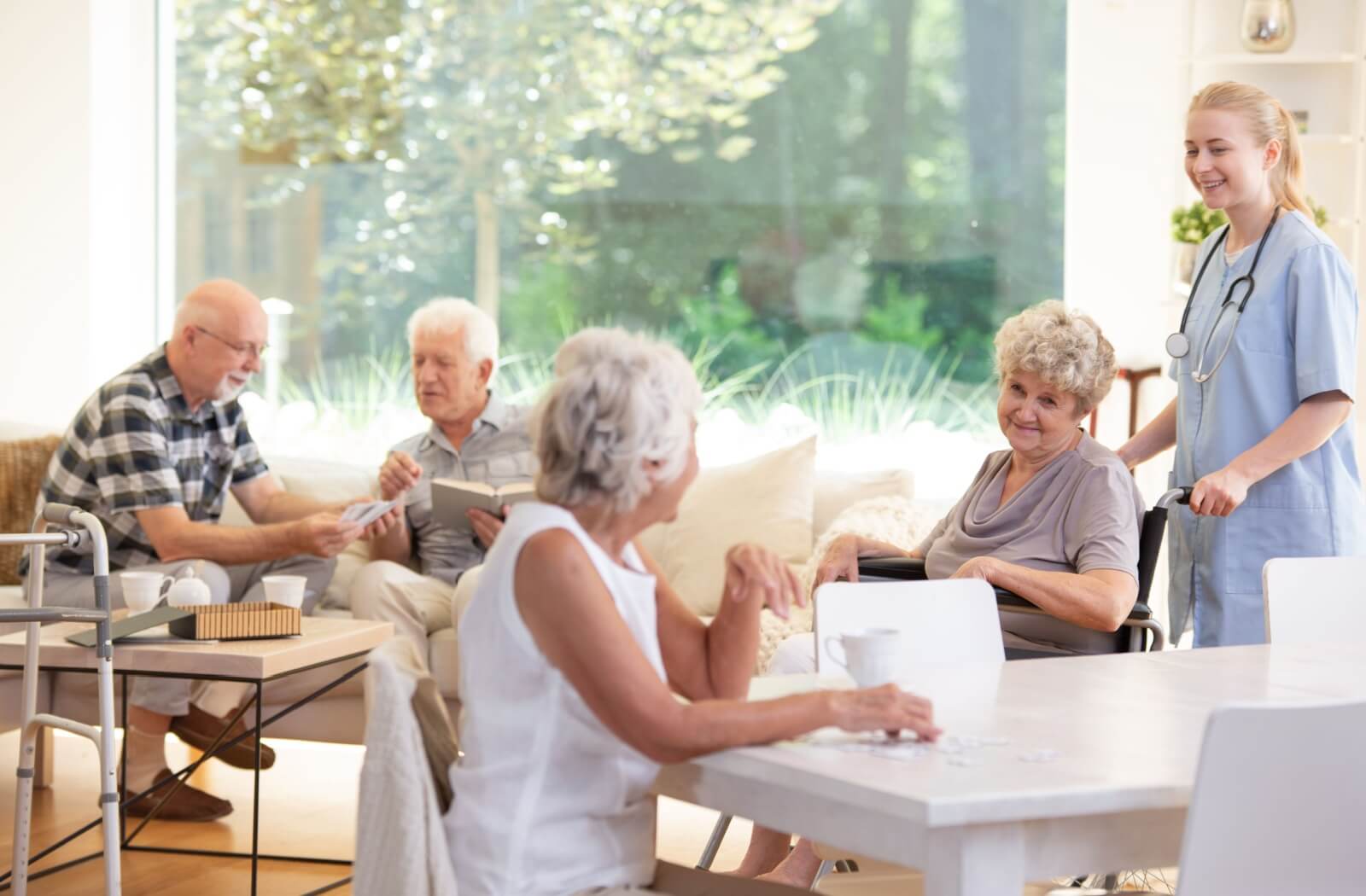 A caregiver checks in on 4 residents in a comfortable sunlit common area in assisted living.