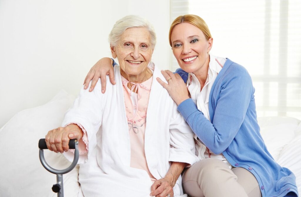 A caregiver and an older adult smile at the camera while sitting on a bed together in assisted living.
